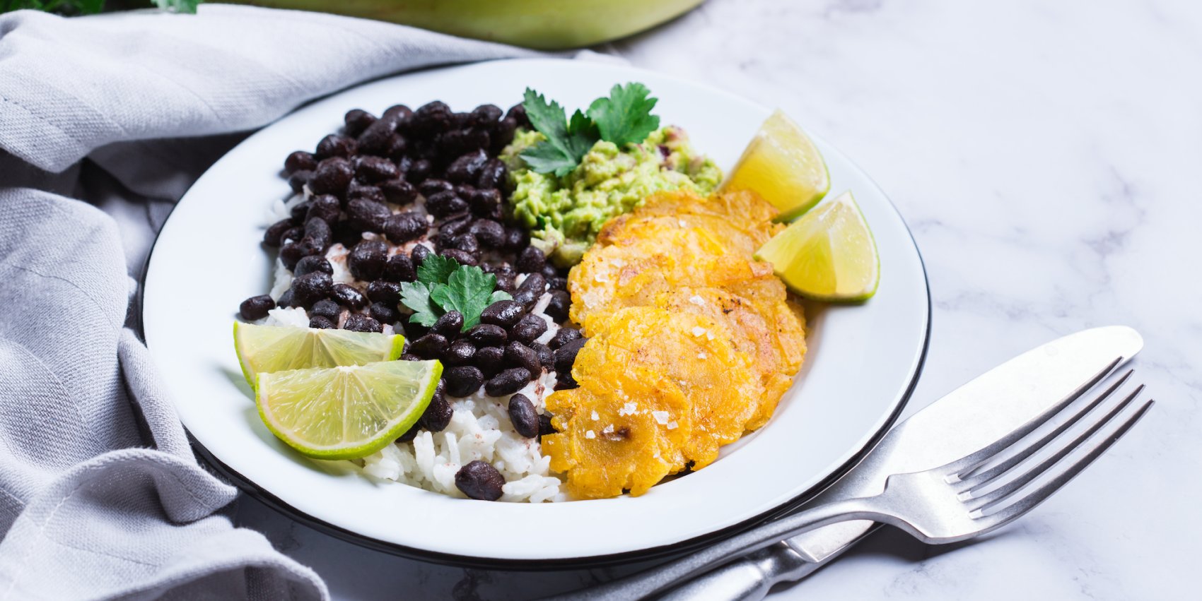 A vegetarian dish consisting of rice, beans, guacamole, plantains, and lime on a white plate