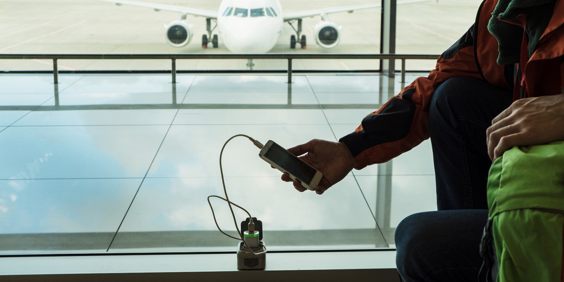 Person sitting at an airport gate charging their phone with a travel adapter with the plane behind them through the windows
