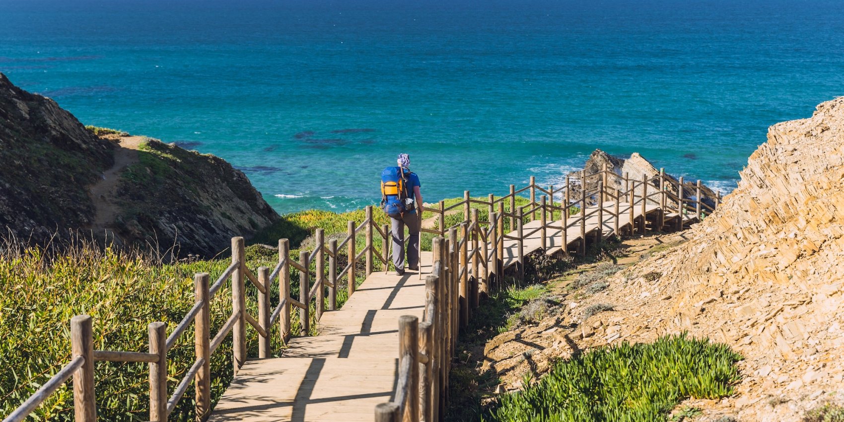Hiker with a backpack walking down a wooden boardwalk on Portugal’s Fishermen’s Trail, surrounded by coastal cliffs and turquoise Atlantic waters.