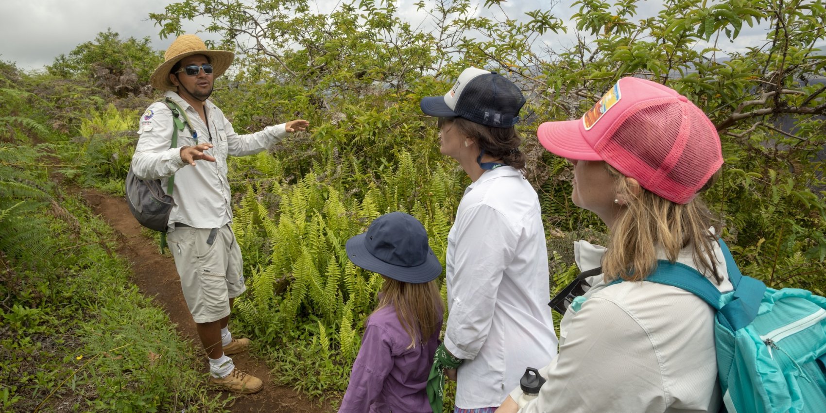 A naturalist guide speaks to a small group of travelers, including a child, along a lush green trail in the Galápagos Islands, highlighting native plants and wildlife.