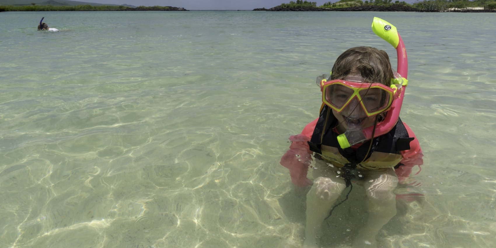 A child wearing snorkeling gear smiles in the shallow, crystal-clear waters of a Galápagos beach, ready to explore the underwater world.