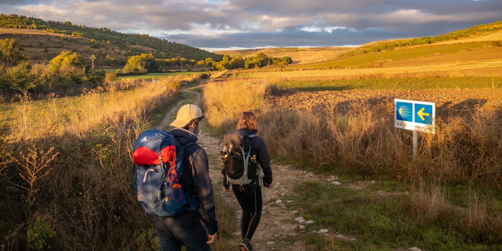 Two hikers walking along the Camino Portugués through the Portuguese countryside, following a marked pilgrimage route at golden hour.