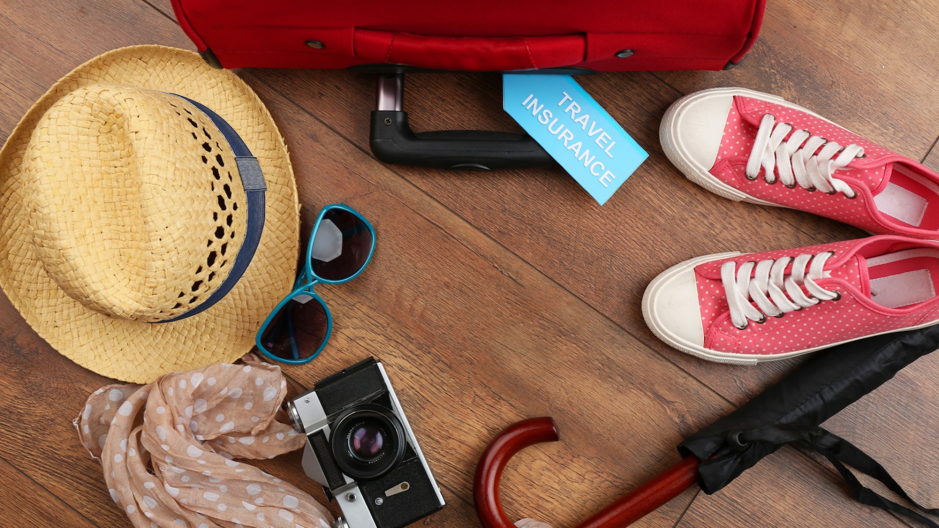 A suitcase, sun hat, sunglasses, camera, scarf, and a pair of shoes laid out on hard wood floors