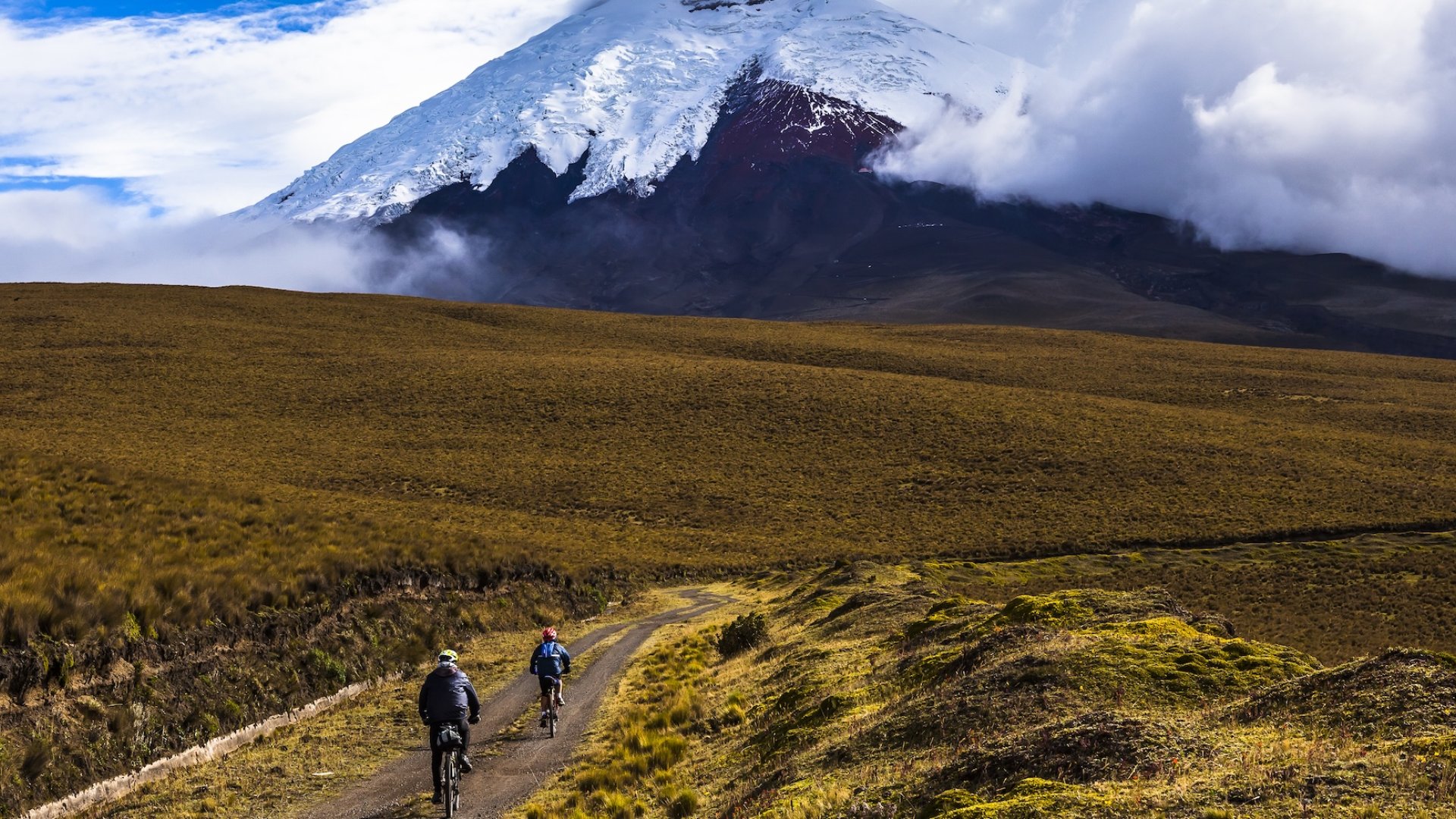 Cyclists riding on a scenic dirt path in the Ecuadorian highlands with Cotopaxi Volcano in the background, a popular mainland extension to Galápagos Islands tours.