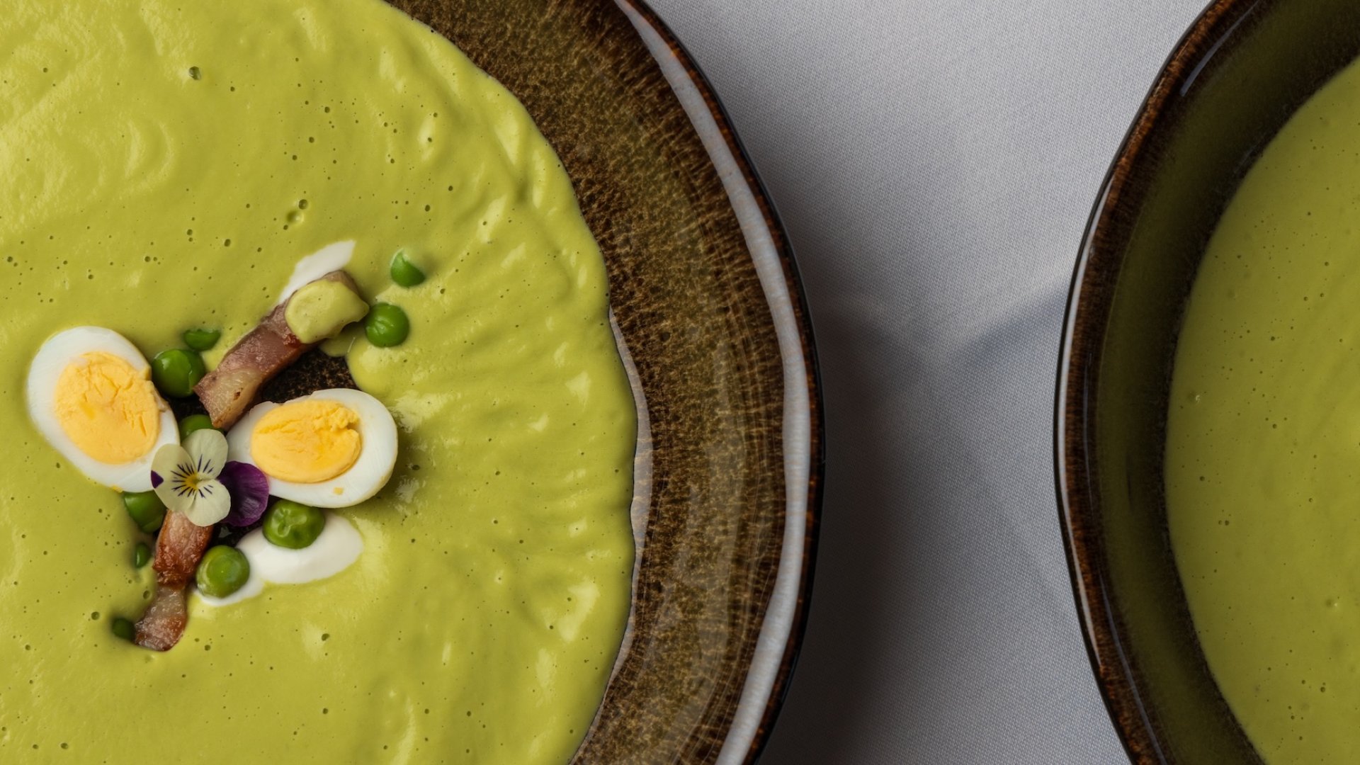 Two bowls of creamy green soup garnished with quail eggs, bacon, peas, and a flower, served in a dark ceramic bowl.