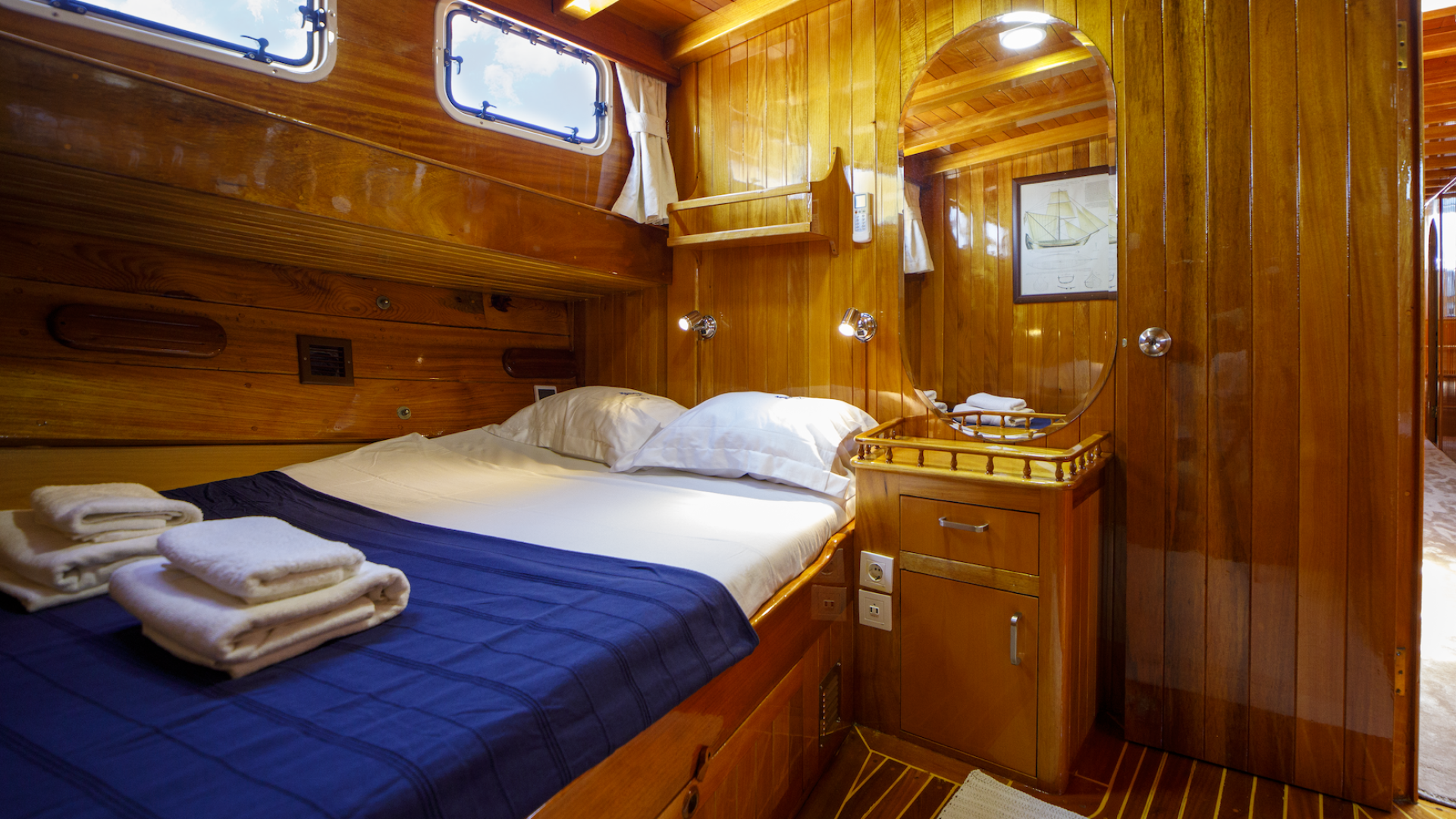 Double bed with white folded towels at the foot, next to a small vanity with a mirror aboard a small yacht in Turkey