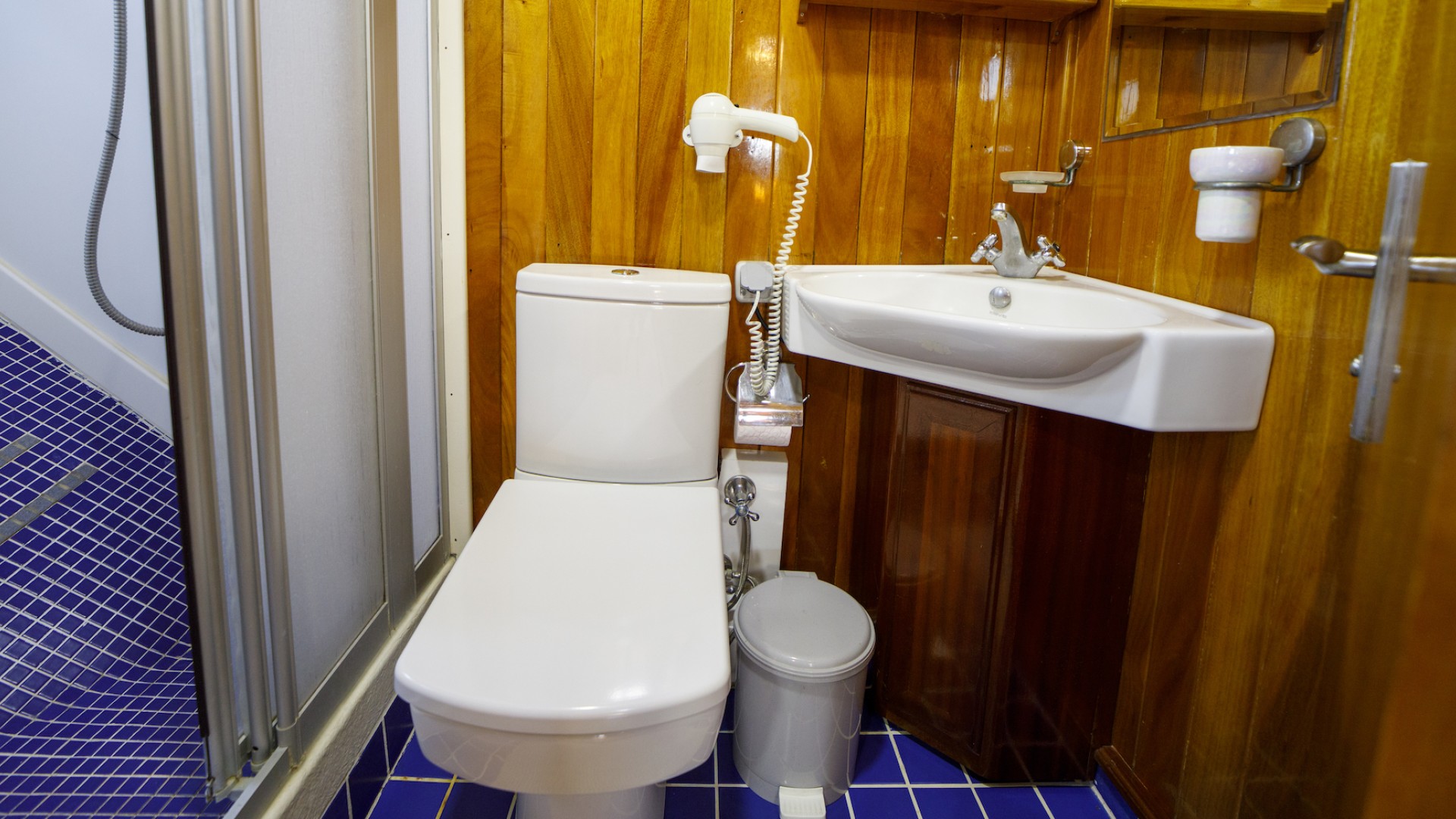 Small bathroom with a white closed toilet, corner sink, mirror on the wall, and shower to the far left