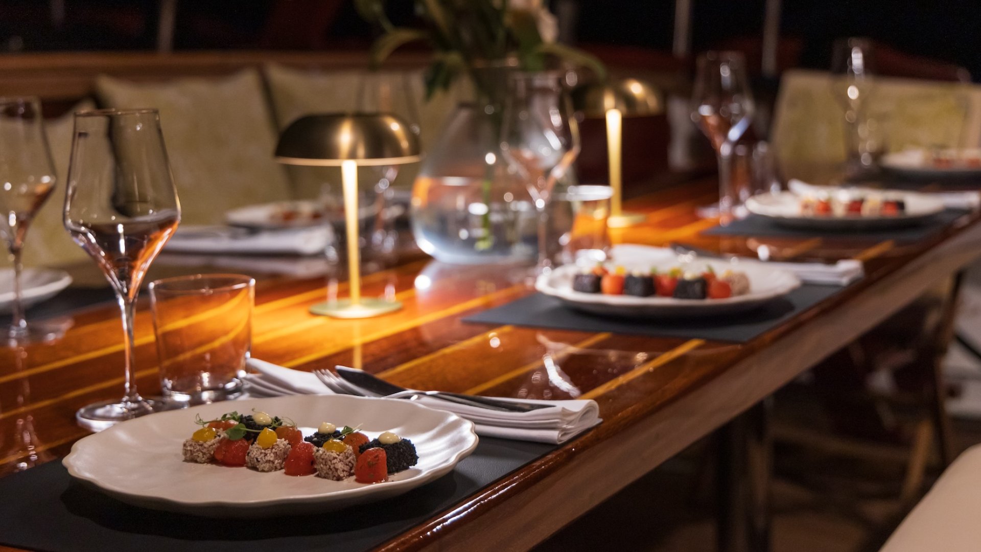 A neatly set dining table on a yacht at night, with gourmet appetizers, wine glasses, and a floral centerpiece.