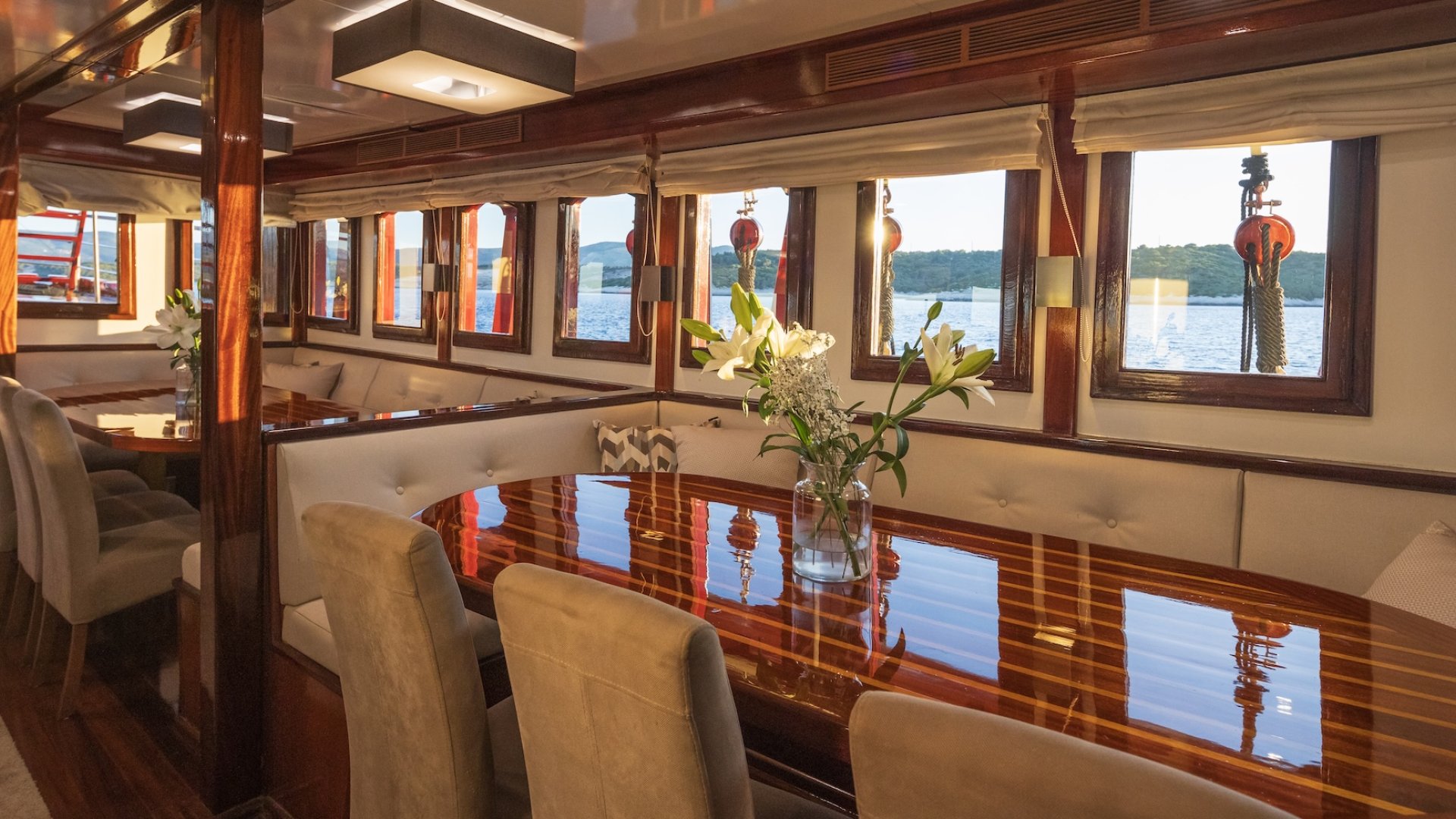 A yacht's indoor dining area with a polished wooden table, cushioned seating, and windows displaying a view of the sea.