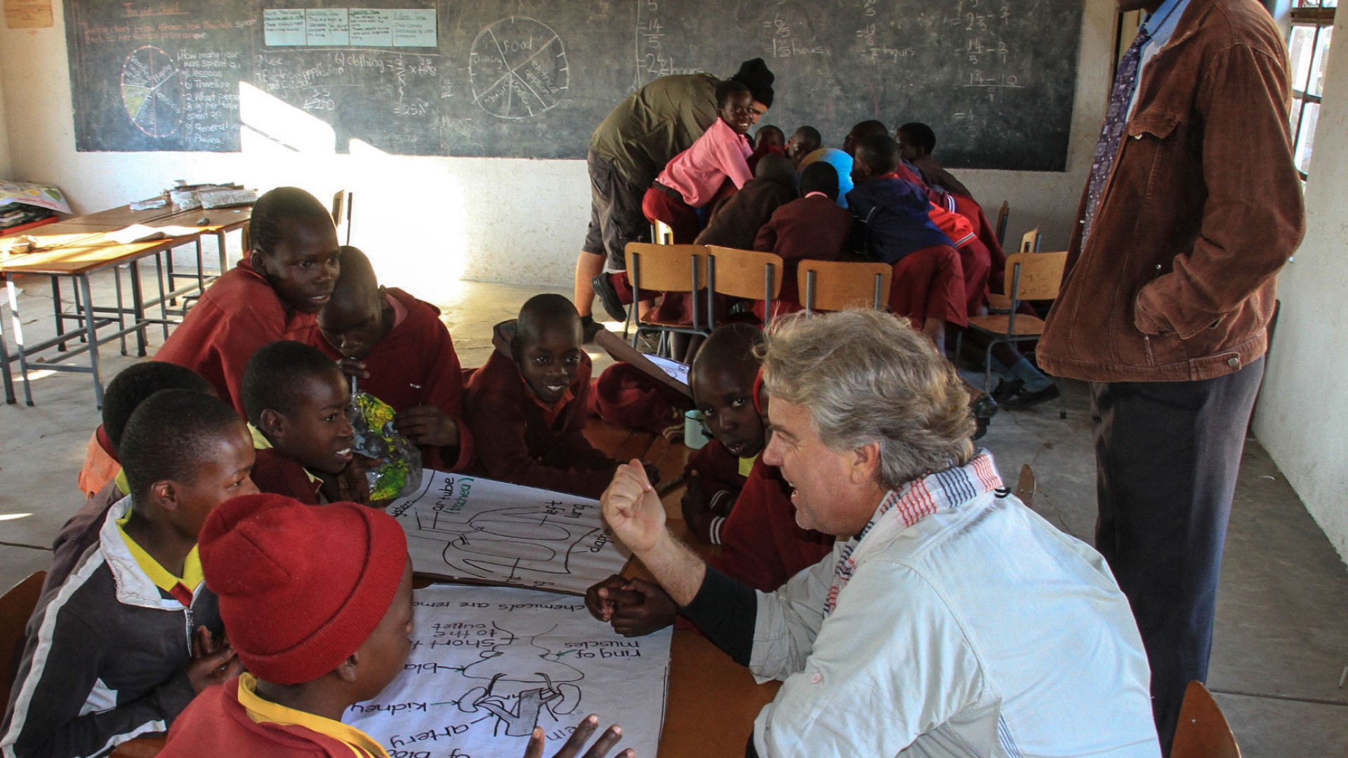 An older man talking to a group of kids around a table with paper on it at a small school in Zimbabwe