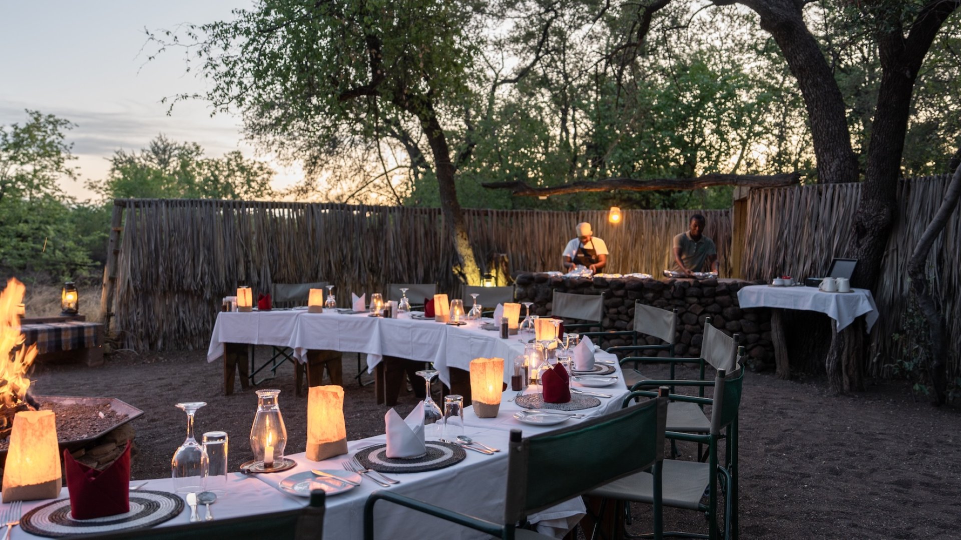 Camp-style outdoor dinner setup with lanterns, firepit, and chefs preparing food in Botswana.