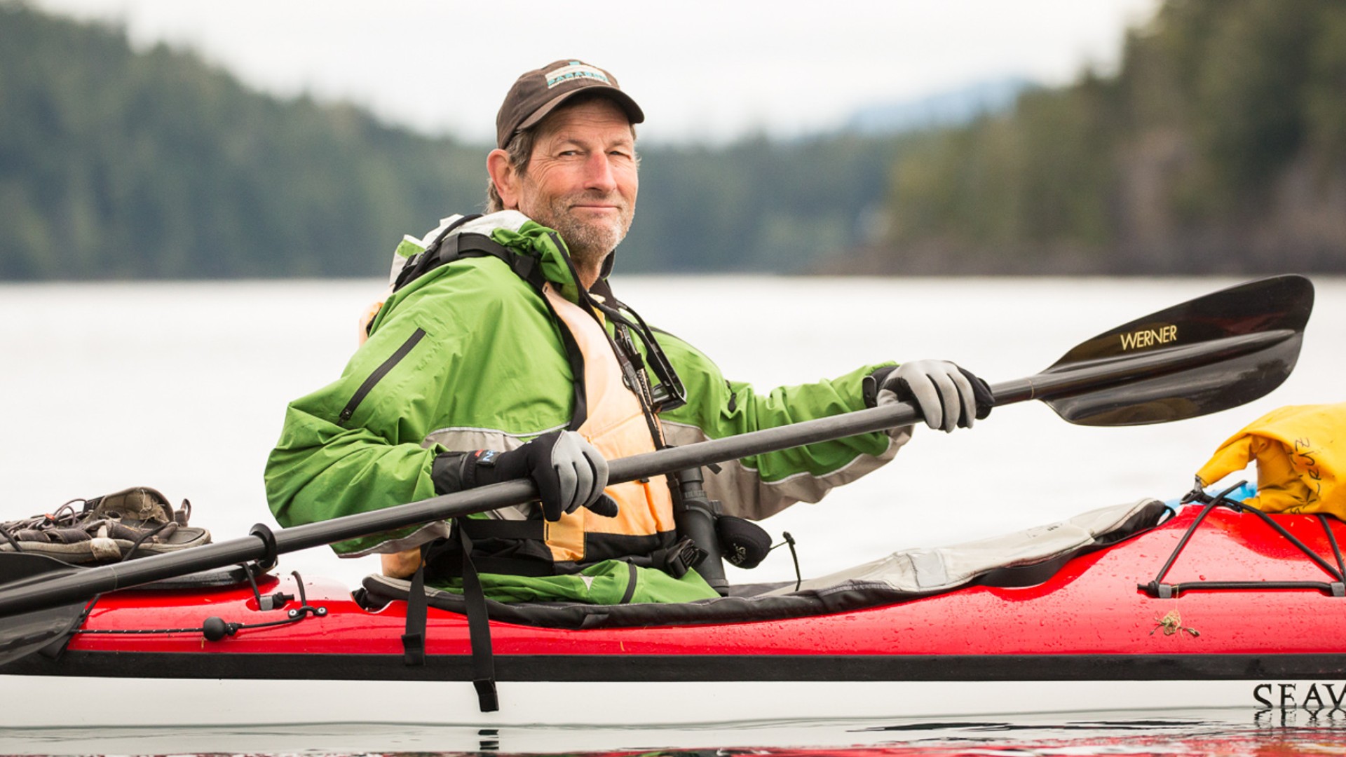 Person in a baseball hat and green jacket in a red kayak smiling while paddling off the coast of Vancouver Island