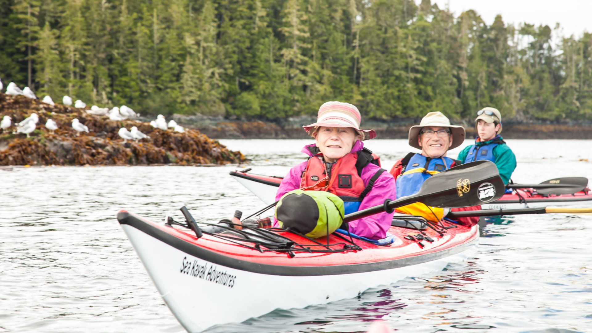 Two people in matching sun hats in a tandem kayak paddle through kelp and seaweed while smiling for the camera