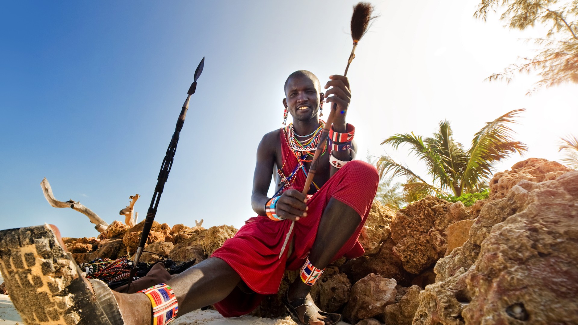 Person of the Maasai tribe in a traditional skirt sitting on the beach in Kenya next to a spear 