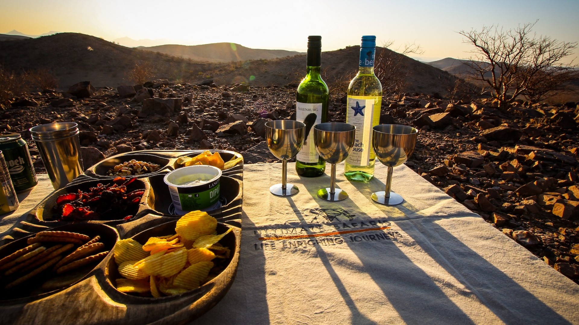 A table with three wine glasses, two bottles of wine, and a spread of chips and other light appetizers with the sunsetting behind it