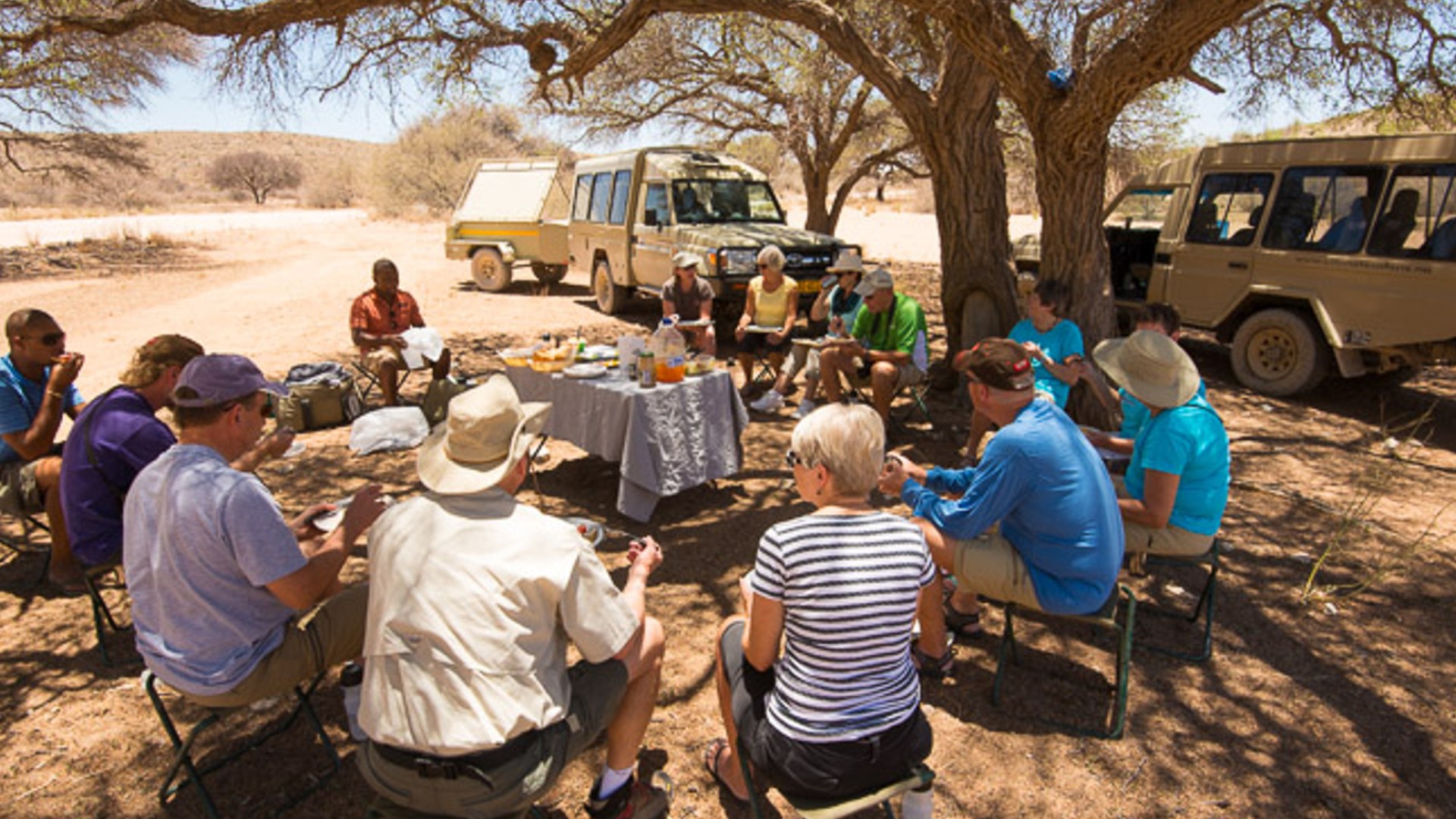 Group of people sitting on camp chair in a circle listening to their safari guide explain something in front of their safari car
