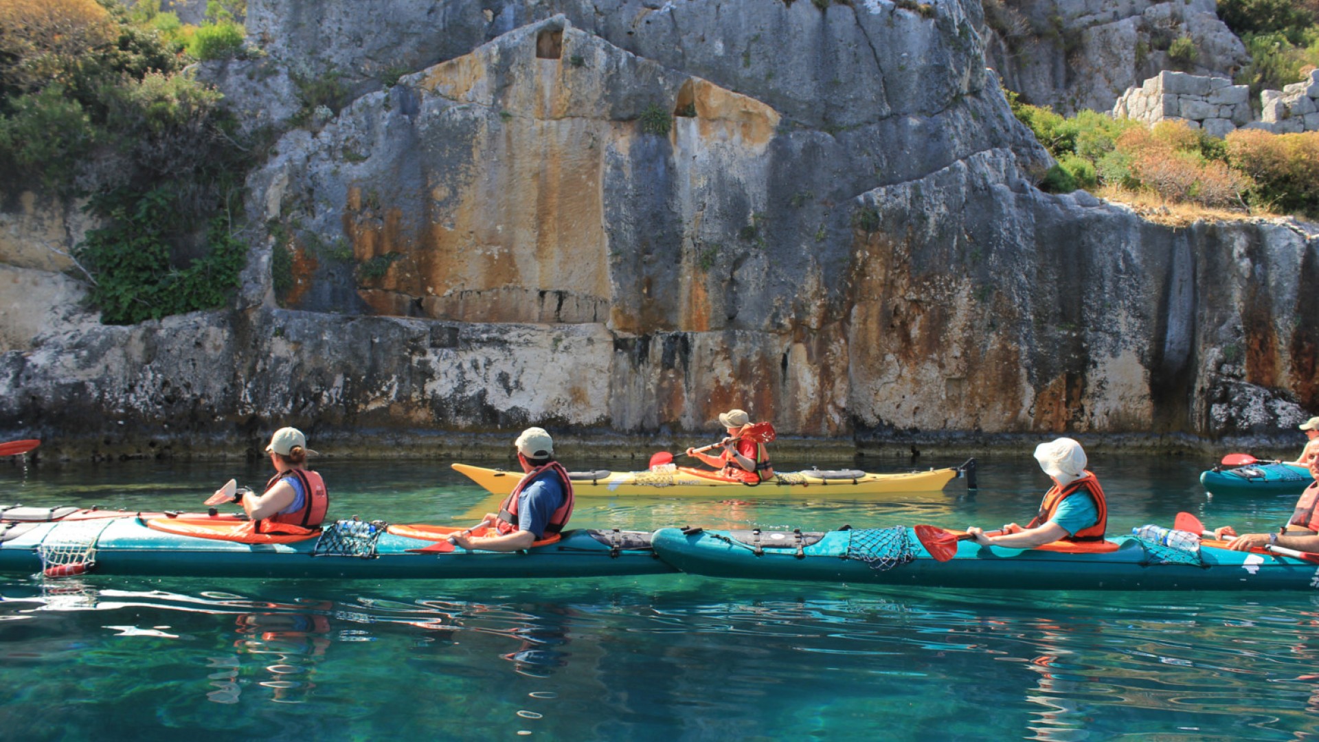 Side view of people sea kayaking atop turquoise waters next to a sheer rock face in Turkey