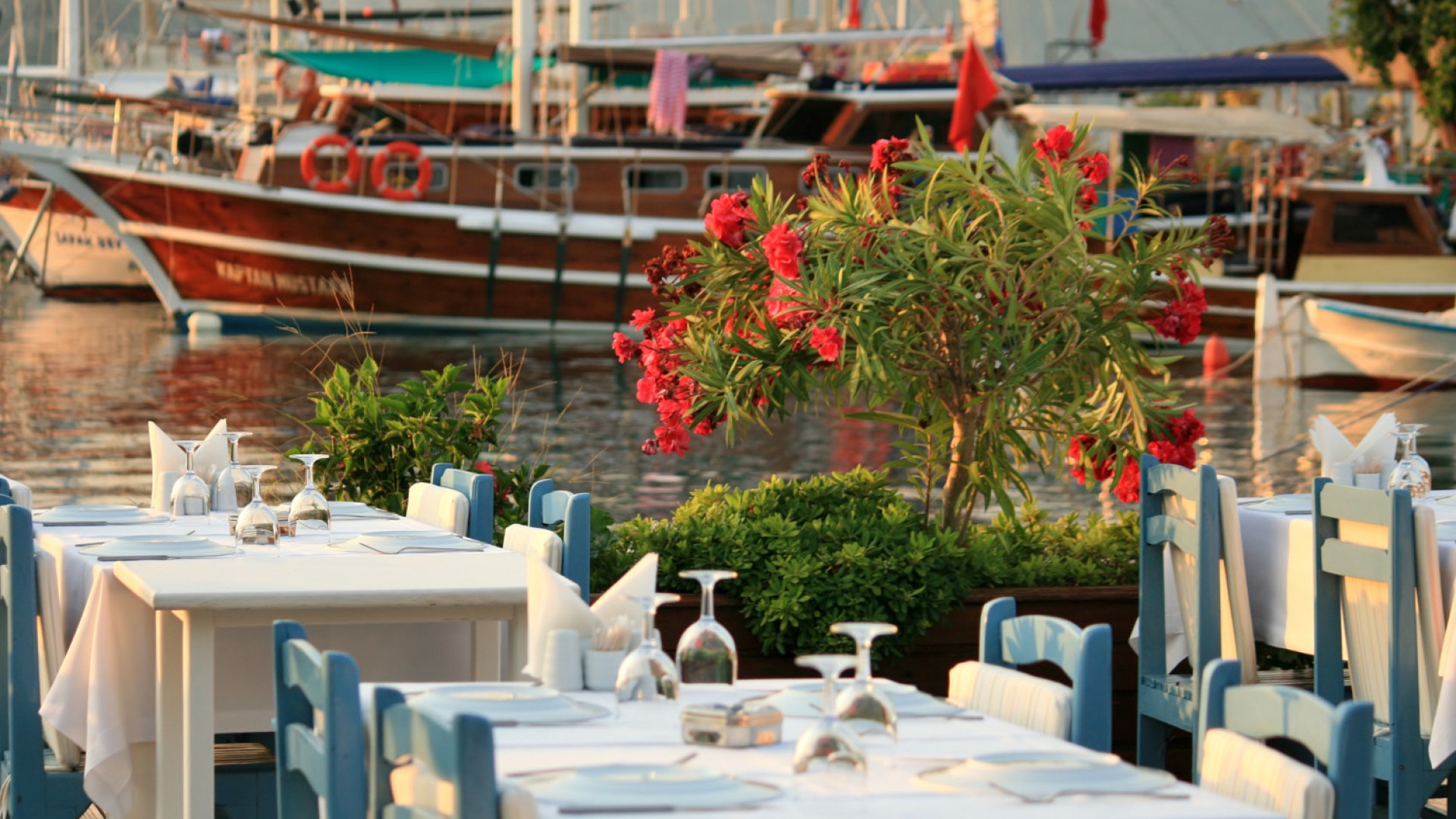 Row of dining tables outside with white table cloths and glassware set up overlooking the marina full of yachts in Turkey