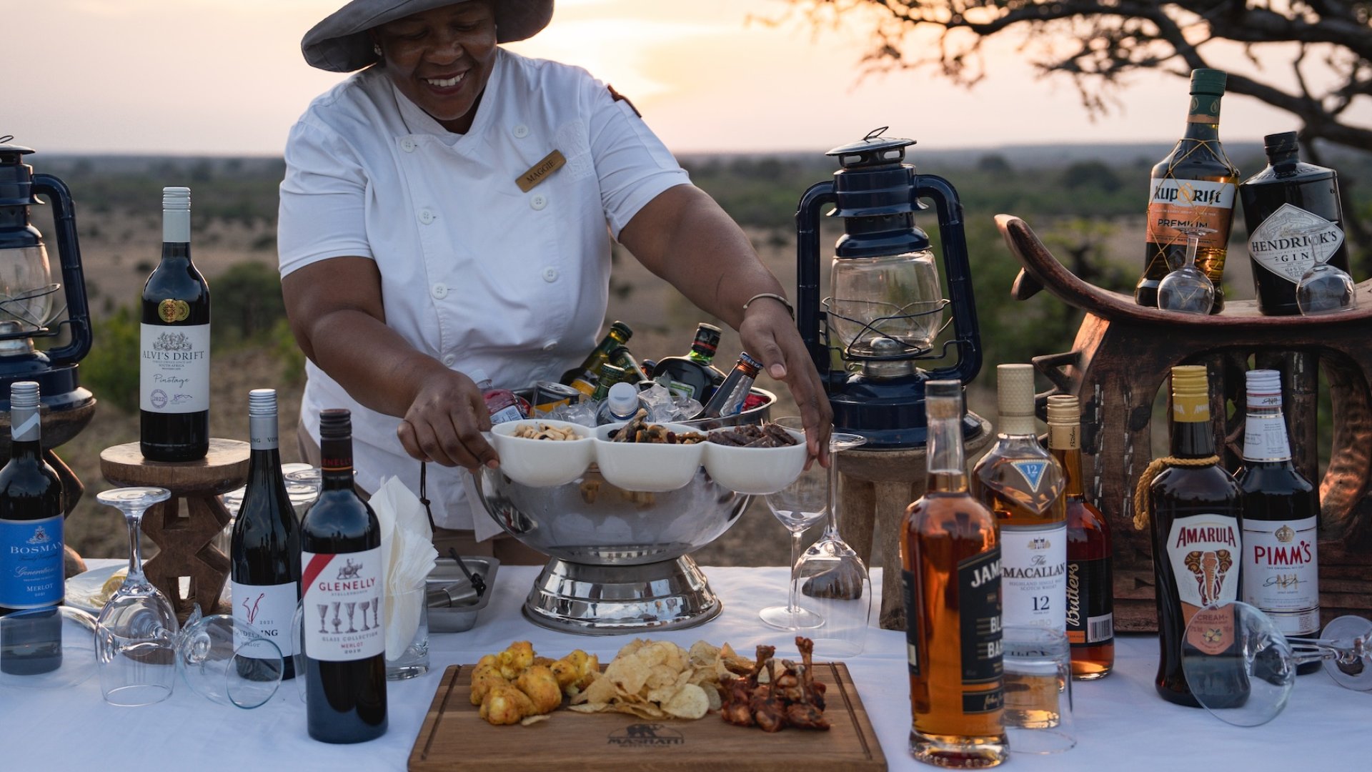 Smiling staff member arranging drinks and snacks at a scenic outdoor bar setup in Botswana.