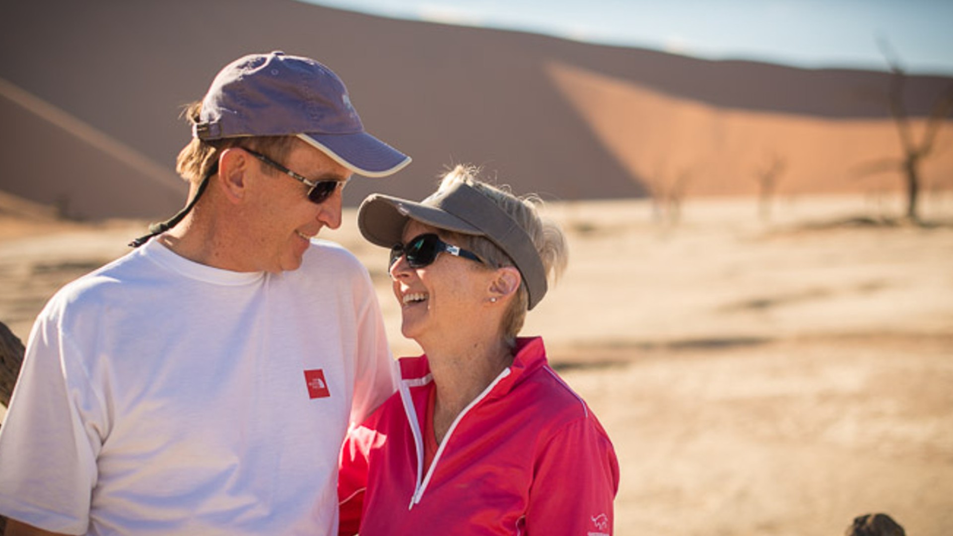 A man and woman both wearing visors standing next to each other and smiling while standing outside their safari car in Namibia
