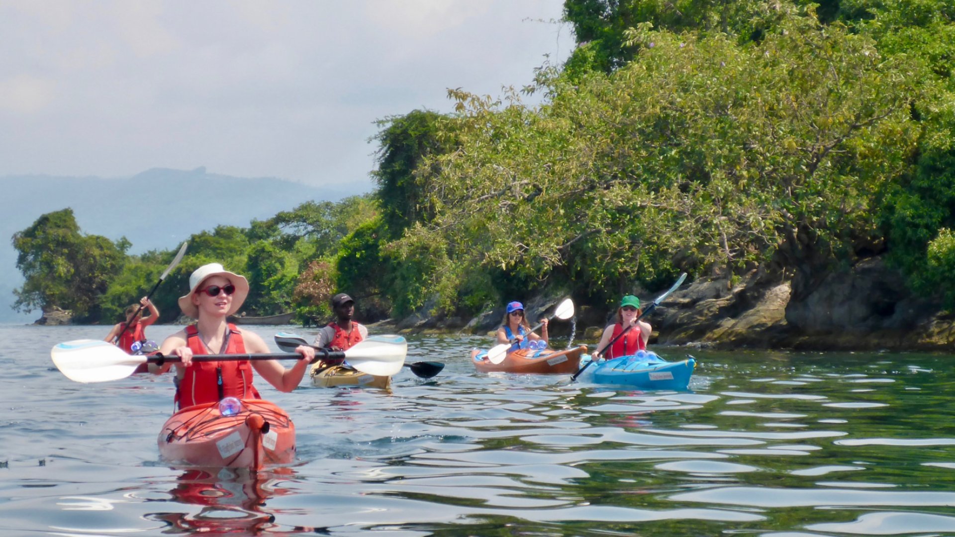 Guided group kayaking near a green, tree-lined shoreline on Lake Kivu in Rwanda.