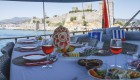 Spread of food and beautifully cut fruit on a table with a white table cloth on a small yacht overlooking Turkish architecture on shore in the distance