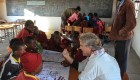 An older man talking to a group of kids around a table with paper on it at a small school in Zimbabwe