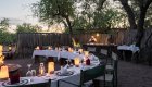 Camp-style outdoor dinner setup with lanterns, firepit, and chefs preparing food in Botswana.