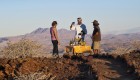 Three people standing around a small table with a yellow table cloth holding appetizers in the middle of the desert in Namibia