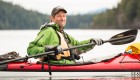 Person in a baseball hat and green jacket in a red kayak smiling while paddling off the coast of Vancouver Island