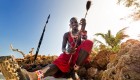 Person of the Maasai tribe in a traditional skirt sitting on the beach in Kenya next to a spear 