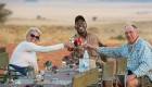 Two tourists with their safari guide at the end of the table holding drinks up to cheers around a table set up on their safari