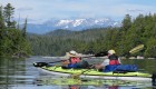 A green tandem kayak with two passengers paddling towards snow capped mountains 