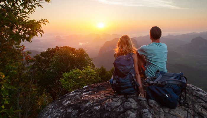 A man and woman sitting on a rock wearing backpacks watching the sunset
