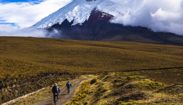 Cyclists riding on a scenic dirt path in the Ecuadorian highlands with Cotopaxi Volcano in the background, a popular mainland extension to Galápagos Islands tours.