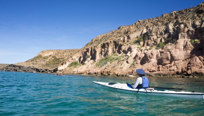 Person in a blue life jacket in a white kayak paddling on the Sea of Cortez past desert rock formations of Loreto Bay National Marine Park
